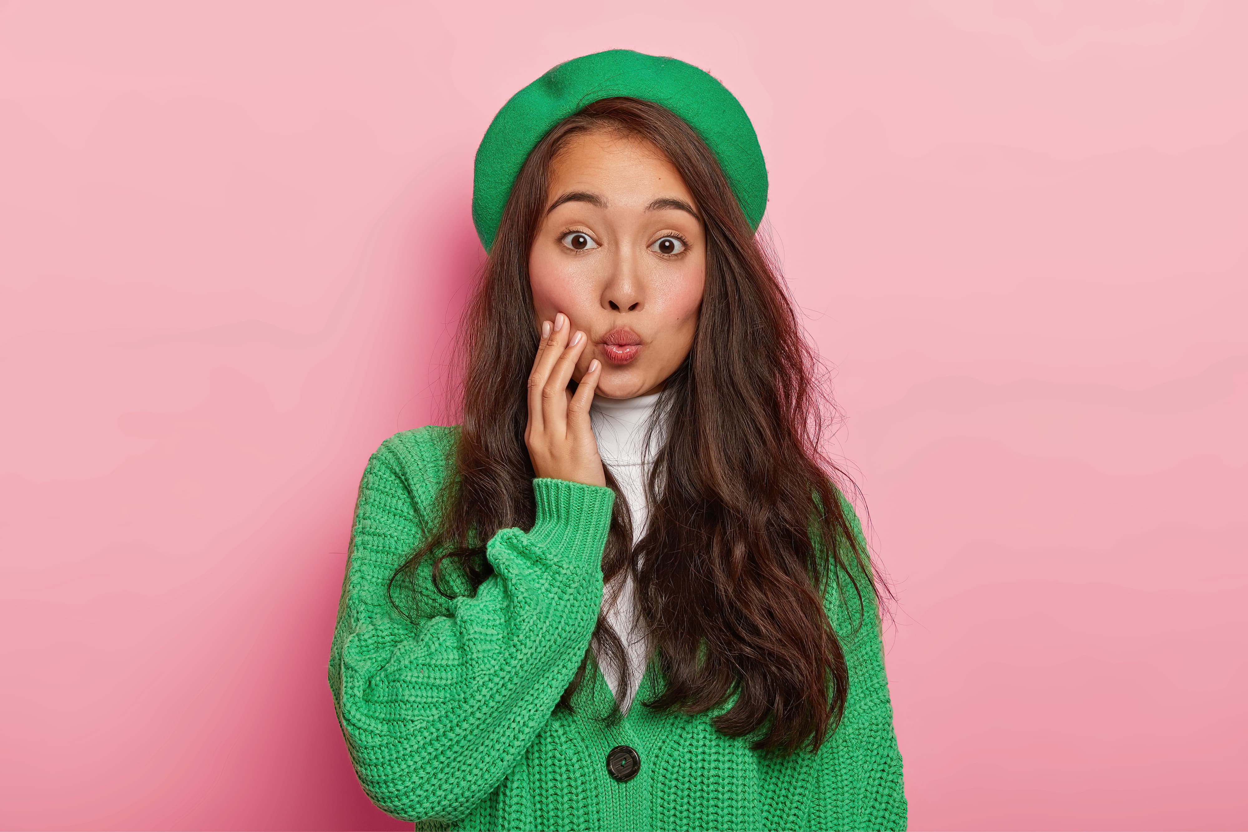 Portrait of good looking Asian lady with dark hair, keeps lips rounded, wants to kiss boyfriend, wears green beret and sweater, poses against pink background
