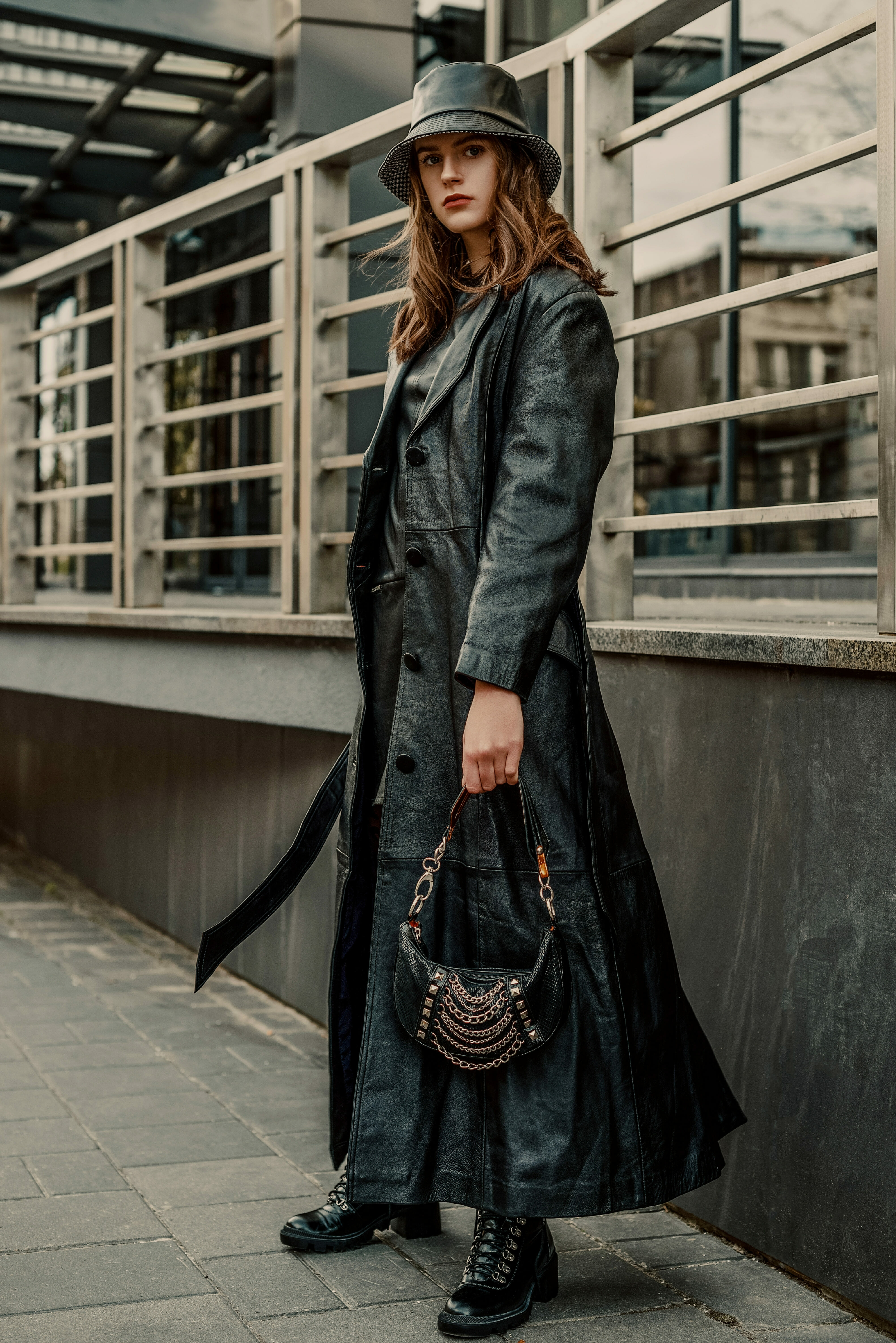 Outdoor full-length fashion portrait of young confident woman wearing long black leather trench coat, bucket hat, lace up ankle boots, holding trendy small handbag, posing in city street
