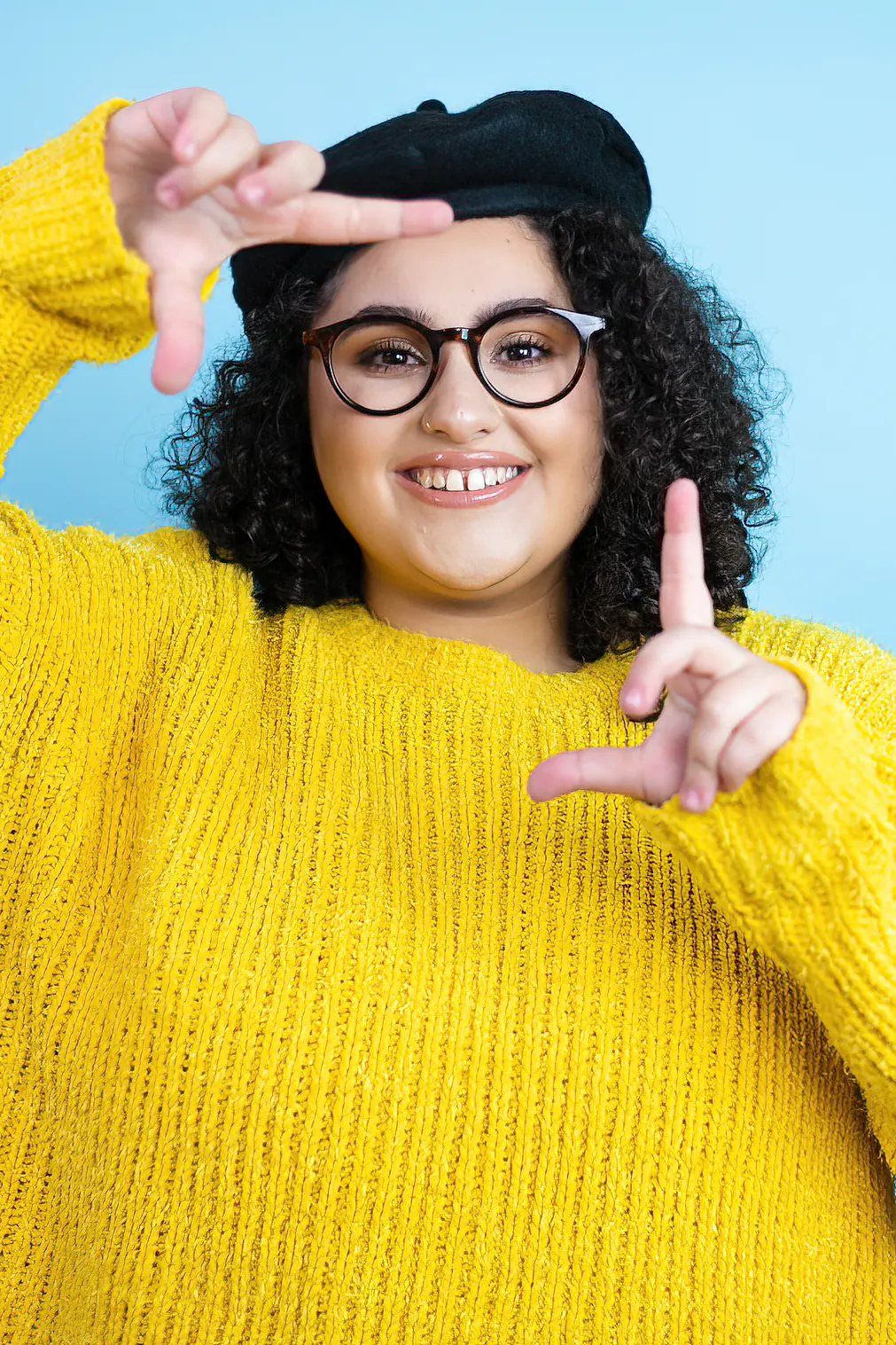 Young beautiful woman wearing black beret and yellow casual sweater over isolated blue background smiling making frame with hands and fingers with happy face