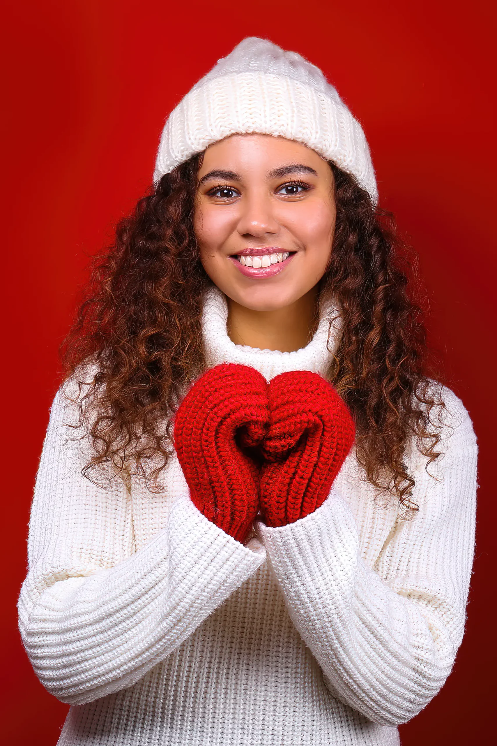 Studio portrait of young woman with dark skin and long curly hair wearing a white beanie knitted turtle neck sweater over the festive red wall