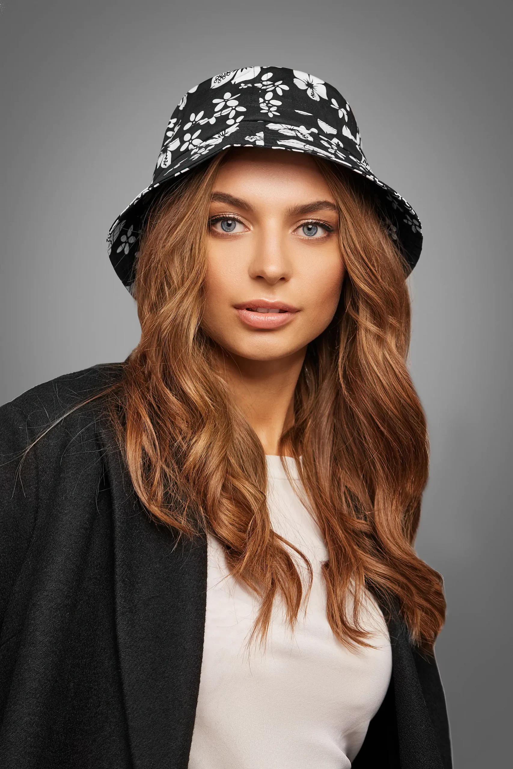 Medium close-up shot of charming blonde lady with wavy hair dressed in a beige top, a black jacket and a black bucket hat with a floral print